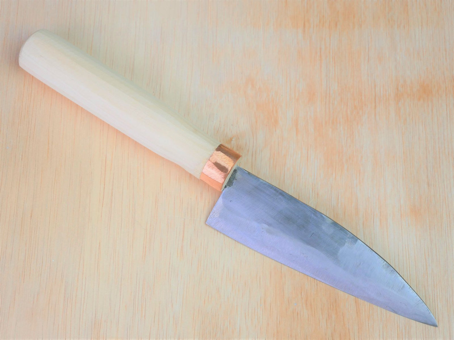 130mm Shirogami No.3 Santoku forged by Tsutomu Takahashi laying on wooden background with it's cutting edge facing south
