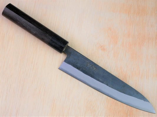 180mm Aogami No.2 Kurouchi Gyuto forged by Yasuaki Taira laying on wooden background with it's cutting edge facing south